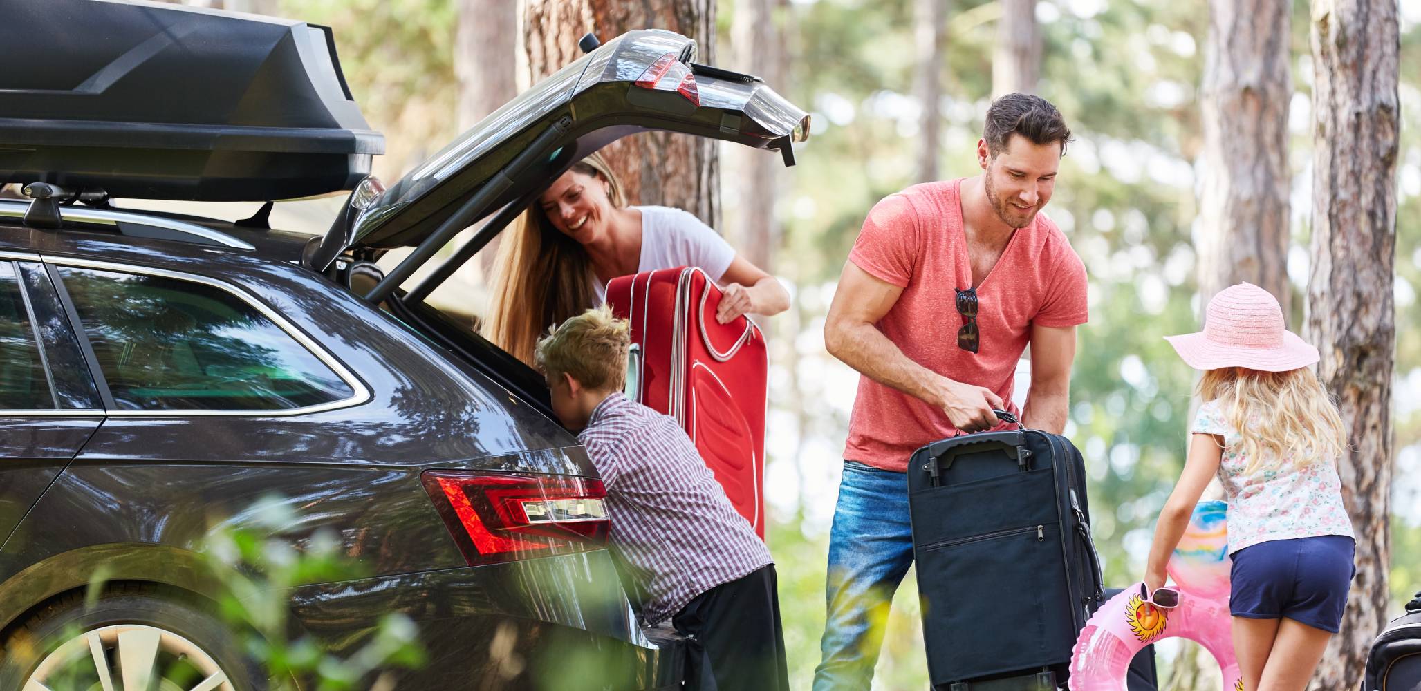 With bright, green woods as a backdrop, a happy family dressed for summer load their luggage into the back of a grey SUV with a roofbox.