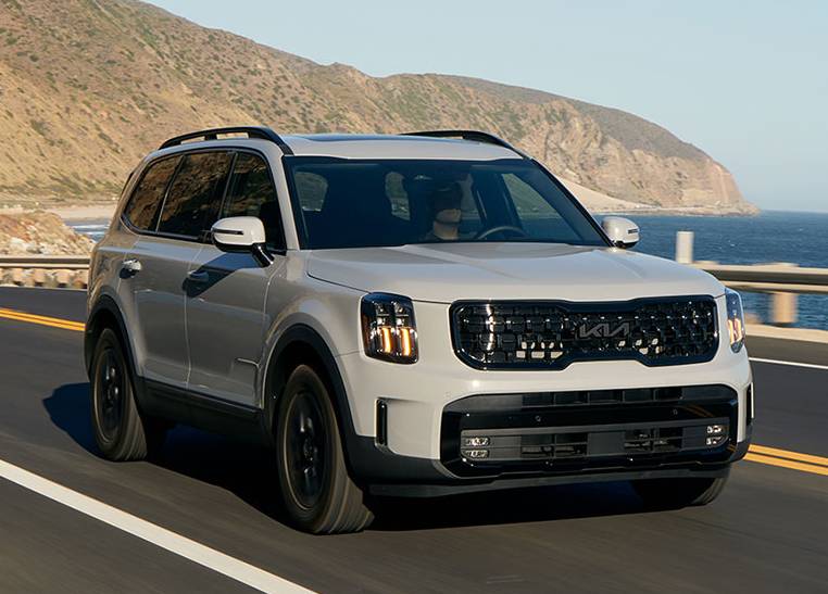 a white 2025 Kia Telluride drives along a highway looking out over the ocean. Sandy cliffs rise in the background.