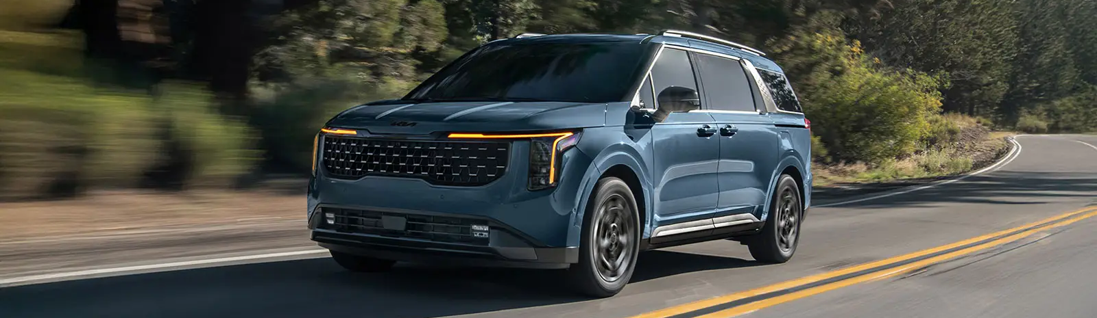 A blue 2026 Kia Carnival drives along a road with greenery in the background