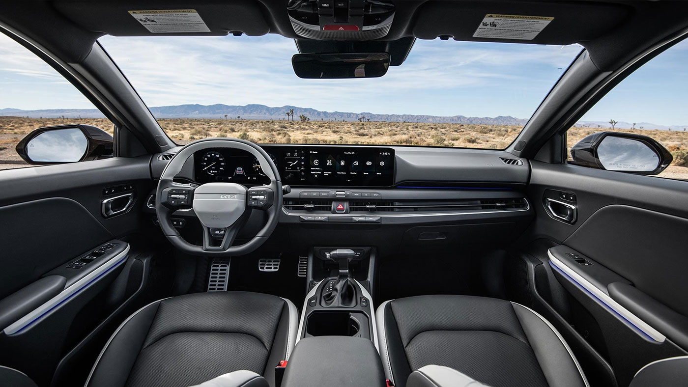 The interior cabin of the 2026 Kia K4 Hatchback, showing the front seats, dashboard, wheel, display, and a horizon with a bright blue sky beyond the windows.