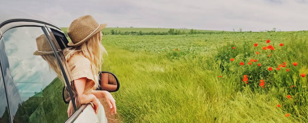 Young woman in a hat enjoying summer vacation. Looking from the window of a car to a green field with poppy flowers. Travel by car.. Kia spring service at Lally Kia in Chatham, ON